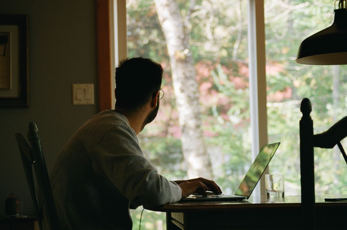 Mann sitzt an einem Esstisch vor seinem Laptop und schaut durch ein gro&szlig;es Fenster in einen Wald.