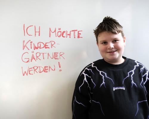 Kind vor einer Tafel, auf der geschrieben steht "Ich will Kindergärtner werden."