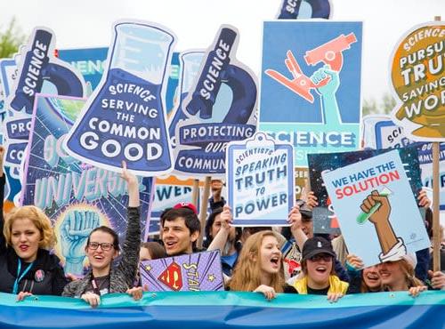 Demonstration von Wissenschaftler*innen. Demonstrant*innen halten Plakate hoch mit der Aufschrift "Science serving the common good" oder "We have the solution".